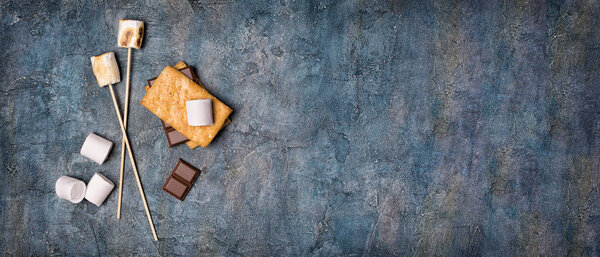 Top view on sticks with roasted on campfire marshmallow, crackers and chocolate as ingredients for s 'mores on blue concrete background with copy space
