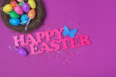 Mutlu Paskalya ve parlak renkli şeker şeker veya dekor mor zemin üzerine kabartma olarak konfeti ile kuş yuvada ışıltılı yumurta pembe metin üstten görünüm