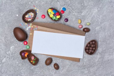 Mockup çikolata geleneksel Paskalya yumurtaları ve parlak renkli badem veya gri beton zemin üzerine konfeti ile boş beyaz kart ve kraft zarfın üzerinde üstten görünüm