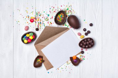 Mockup çikolata geleneksel Paskalya yumurtaları ve parlak renkli badem beyaz ahşap zemin üzerinde beyaz boş kart ve kraft zarfın üzerinde üstten görünüm