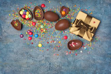 Çikolata geleneksel Paskalya yumurtaları ve altın hediye kutusu ile parlak renkli badem, şeker sprinkles kopya alanı mavi beton zemin üzerine üstten görünüm