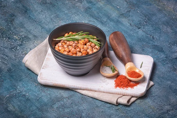 Roasted spicy chickpeas in black bowl with rosemary, sea salt and red pepper in wooden spoons — Stock Photo, Image