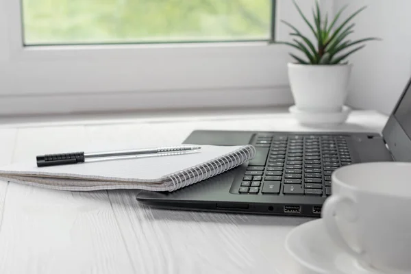 Modern workplace for distance learning at home with black laptop, pen and notebook for writing on light window background