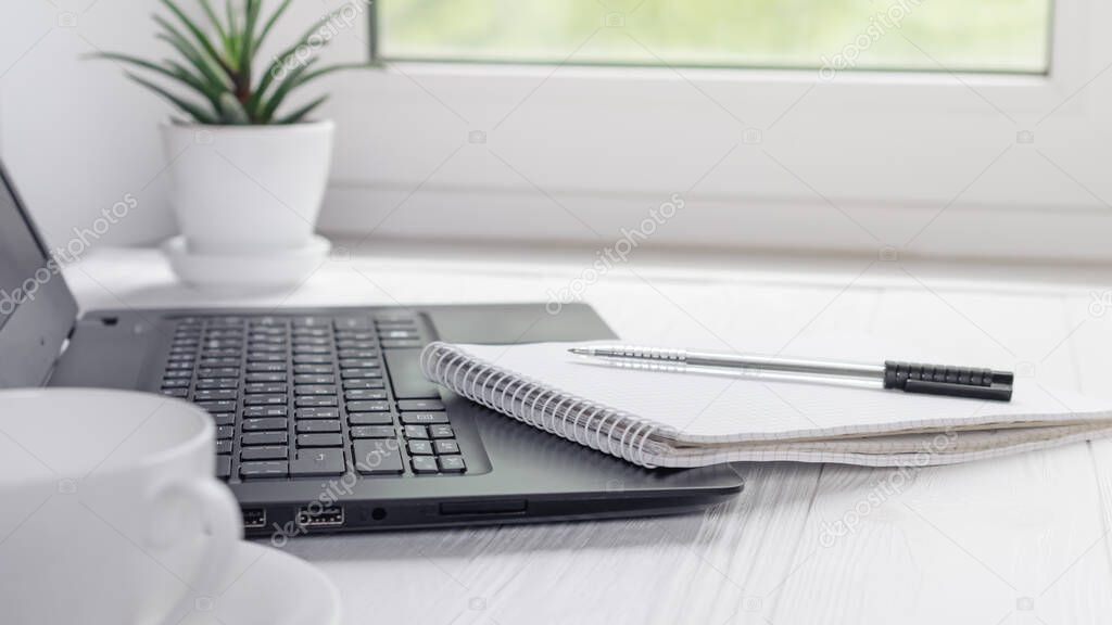 Modern workplace for distance learning at home with black laptop, pen and notebook for writing on light window background
