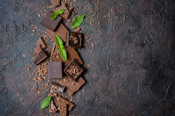 Kırık süt ve koyu çikolata parçalarının üst görüntüsü. Kahverengi beton zemin üzerinde taze yeşil nane ve fotokopi alanı var.