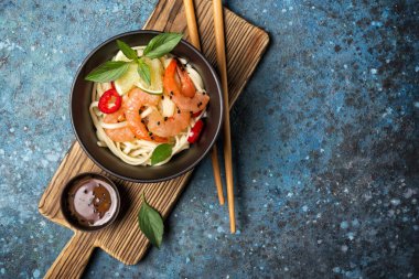 Japon erişteleri, baharatlı karidesler, turuncu Asya sosu, ahşap tahtada limon ve siyah susam tohumları ve kopyalanmış mavi beton arka plan.