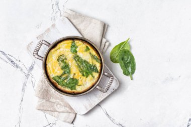 Yeşil ıspanak yapraklı lezzetli omletin üst görüntüsü tavada, ahşap tahtada keten peçete ve fotokopi alanı olan beyaz mermer arka plan.