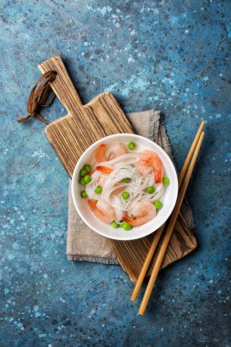 Çin pilavlı erişte, lezzetli karides, yeşil bezelye ve ahşap tahtada ahşap çubuklar ve mavi beton arka plan.