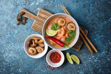 Tom Yam domates çorbası, lezzetli karidesler, midyeler, şiitake mantarları, limon, acı biber, ahşap çubuklar ve mavi beton arka plan.