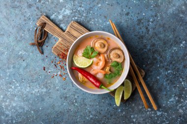 Tom Yam domates çorbası, lezzetli karidesler, midyeler, şiitake mantarları, kireç, biber ve ahşap çubuklar ahşap tahtada ve mavi beton arka planda.