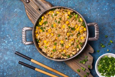 Tavada tava, Çin pirinci, tavuk yumurtası, sarı mısır ve yeşil soğan ahşap tahtada ve mavi beton arka planda.