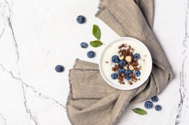 Ev yapımı yoğurt, yulaf gevreği veya granola, fındık, taze yaban mersini ve yeşil nane yaprakları beyaz mermer arka planda gri keten peçeteyle.
