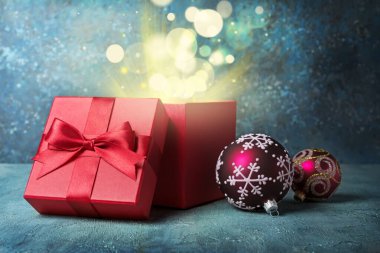 Parlak saten fiyonklu güzel kırmızı kare hediye kutusu şimdiki ve sihirli ışık için, mavi beton arka planda Noel ağacı için oyuncaklar