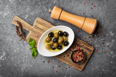 Lezzetli karışık yeşil ve siyah zeytin fesleğen yapraklı ahşap biber değirmeni gri beton arka planda.