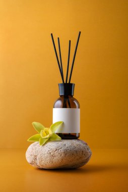 Sarı arka planda vanilyalı orkide çiçeği ve boş etiketli kehribar cam kamış ayırıcı. Markalaşma, lüks spa, sağlık ve organik tasarım için minimalist aromaterapi ve parfüm modeli.