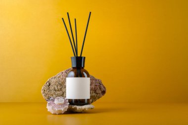 Sarı arka planda deniz kabuğu dekoru olan taşın üzerindeki boş etiketli kehribar cam difüzyon şişesinin bir kopyası. Markalaşma, paketleme ve kozmetik tasarım için doğal eko tarzı şablonu.