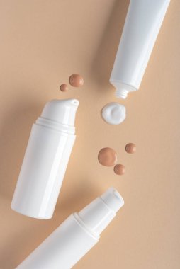 Beyaz tüpler, krem damlaları ve bej arka planda fondöten ile minimal kozmetik kompozisyon. Cilt bakımı, güzellik, makyaj ve sağlık kavramları için mükemmel..