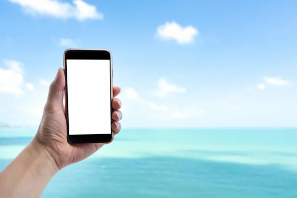 Close up man hand holding black smartphone on beautiful calm blue sea and white sky background. Male hand using blank white screen mobile phone in outdoor summer vacation to taking picture.