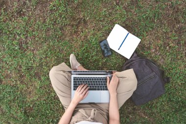Üstten görünüm bahçesinde bulunan smartphone, çantası, defter ve kalem çalışmak için dizüstü bilgisayar kullanan genç serbest meslek sahibi kadın. Mutlu kız blogger blog içeriği park açık yazıyor. Yaşam tarzı çalışma ve aygıt.