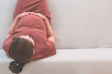 Gri kanepe ve kopya alanı ile üst görünümünde karnı tutan el üzerinde oturan genç güzel hamile kadın. Gebelik, aşk, sağlık, annelik, insanlar, gevşeme ve beklenti kavramı.