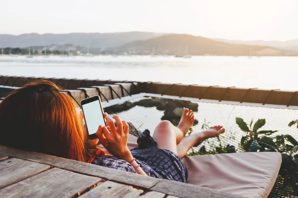 Young woman using black smartphone with blank desktop screen while lying side the sea with evening sunset background. Mockup image of woman hand holding phone chatting online message with friends.
