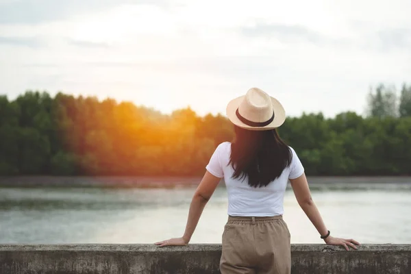 在黄昏时分 快乐的时髦女人戴着帽子站着看河 年轻的亚洲游客享受在大自然的户外湖泊的时间 生活方式旅行 放松和自由概念 — 图库照片