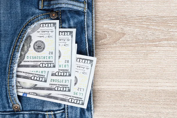 Money in pocket of blue jean on wooden background with copy space. One hundred dollar bills in back of jean pocket.