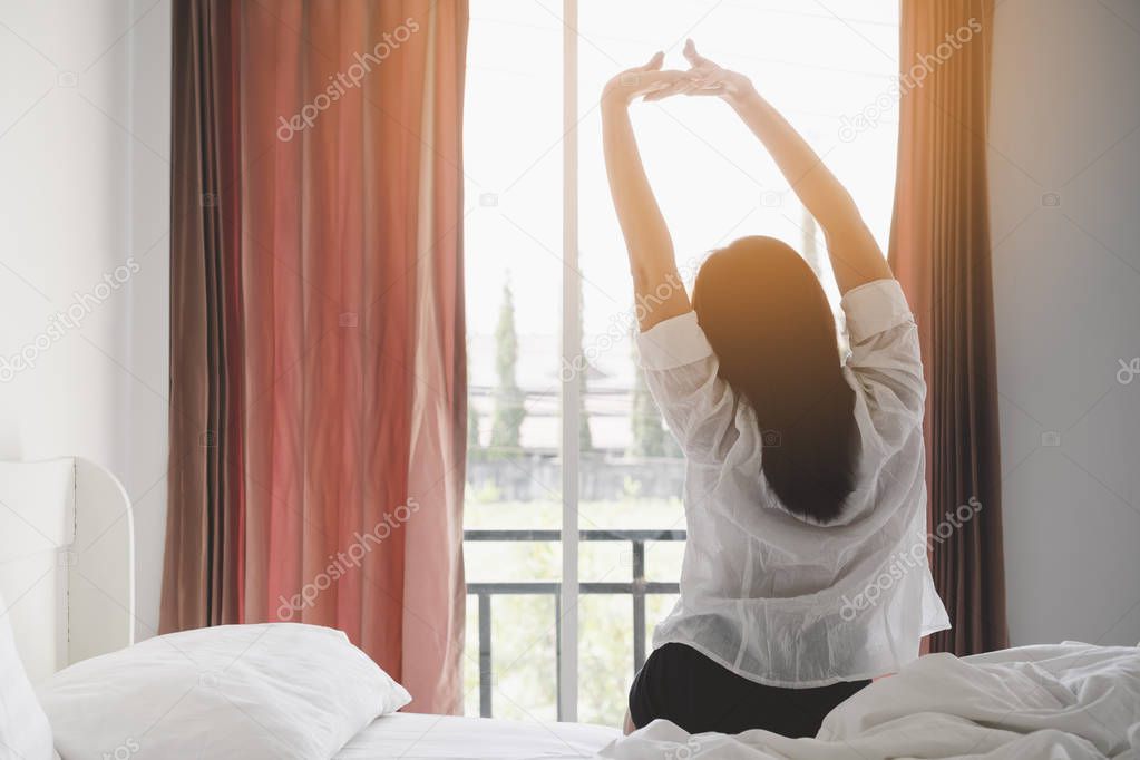 Happy asian woman stretching on bed after waking up at morning in bedroom with sunlight shine through curtains