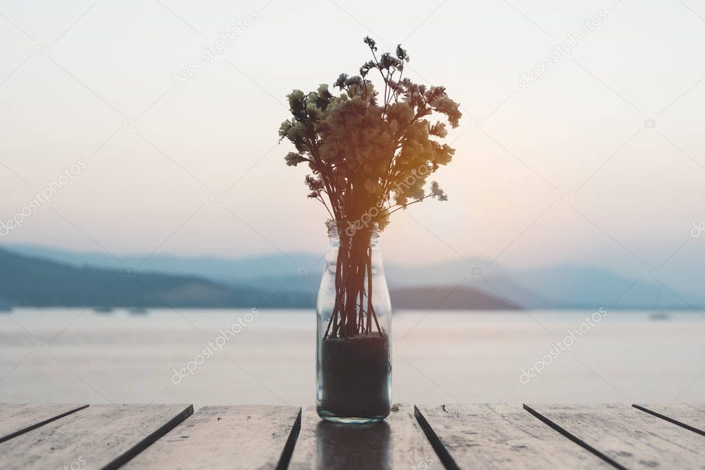 Bouquet of dried flowers in glass jar or bottle on wooden table over blurred sunset behind mountain and sea view background. Vase on table outdoor decoration with dramatic evening dawn scenic.
