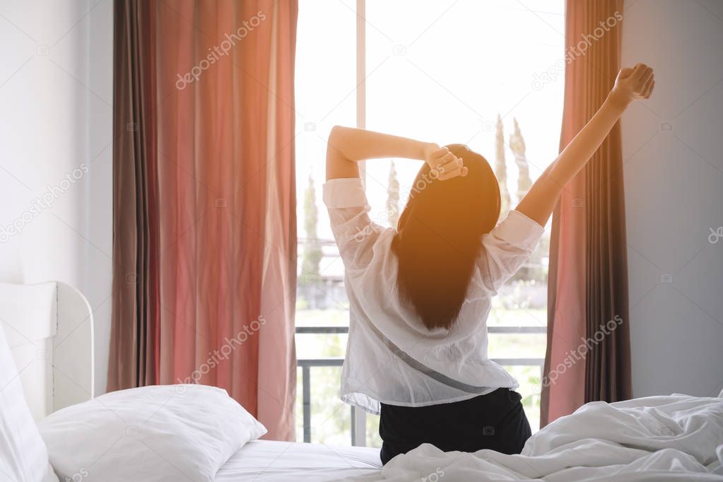 Happy asian woman stretching on bed after waking up at morning in bedroom with sunlight shine through curtains