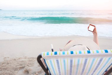 Arka görünümü mutlu kadın plaj sandalyesini boş beyaz ekran smartphone fotoğraf tatil tropikal kumsalda alarak tutan oturan şapkalı. Cep telefonu yaz mavi deniz kullanarak erkek. Yaşam tarzı, mockup kavramı.