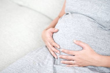 Gri kanepe evde ve karnı dokunmadan el üzerinde oturan mutlu güzel hamile kadın. Mutlu hamile kadın aşk ve annelik kavramı içinde kanepede rahatlatıcı. Gebelik sağlıklı ve gevşeme.