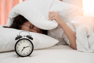 Close up alarm clock with young asian woman lazy waking up in early morning for routine daily job. Sleepy girl lying on bed closing her eyes and try to hiding under pillow for  snooze alarm clock. clipart