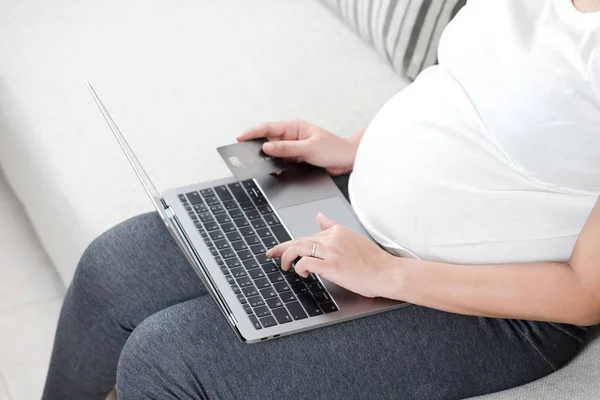 Close up happy young pregnant woman using laptop for shopping online with credit card payment. Smart and modern mom lifestyle sitting on sofa with blank computer screen and hand holding credit card.