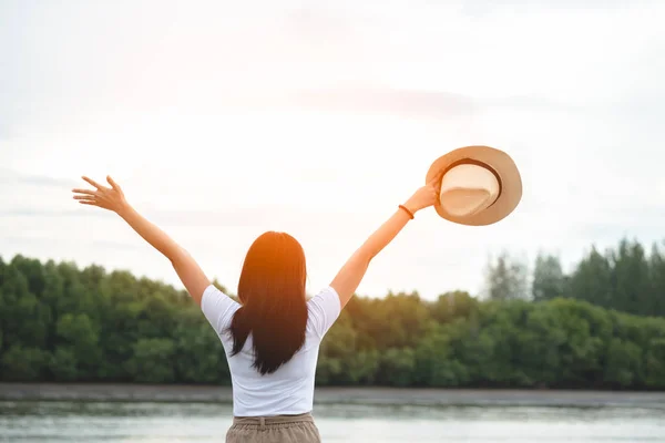 Happy hipster woman wear white shirt standing outdoor open hand and holding hat at lake in evening sunset. Cheerful young girl enjoying life time. Lifestyle travel, relaxation and freedom concept.