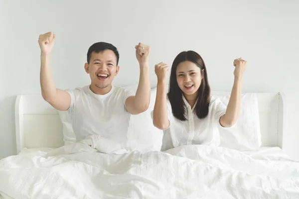 Young couple sport fans cheering for their team victory while watching football match on tv at home. Portrait of asian couple in white casual have fun and celebrating on white bed enjoying together.