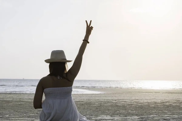 在日落时 坐在热带海和海滩上的快乐女人的背面显示两个手指或胜利标志 年轻女子享受生活假期和放松在天堂岛的复古色调 — 图库照片