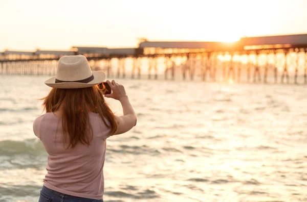 年轻的亚洲妇女在帽子是采取日落海滩照片与智能手机 旅行者女孩拍摄的视频美丽的日落景观在手机相机 暑假旅游和度假 — 图库照片