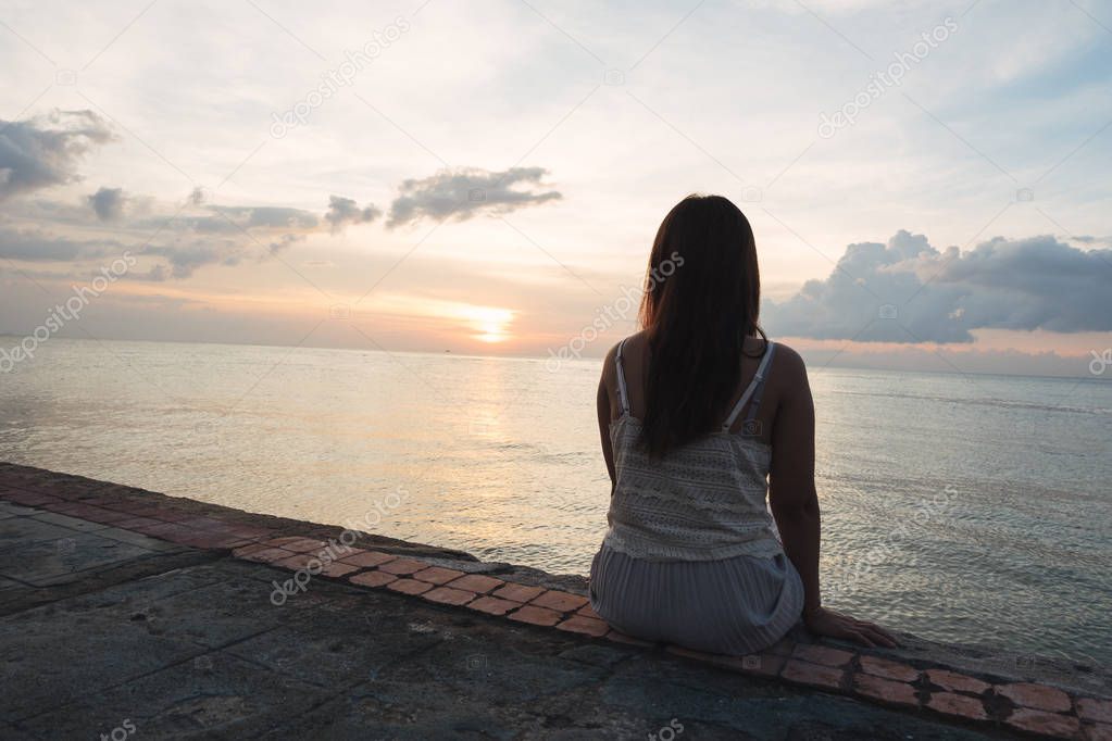 Silhouette of young woman sitting alone on back side outdoor at tropical island beach missing boyfriend and family in summer sunset. Sad and lonely concept in dark and vintage tone.