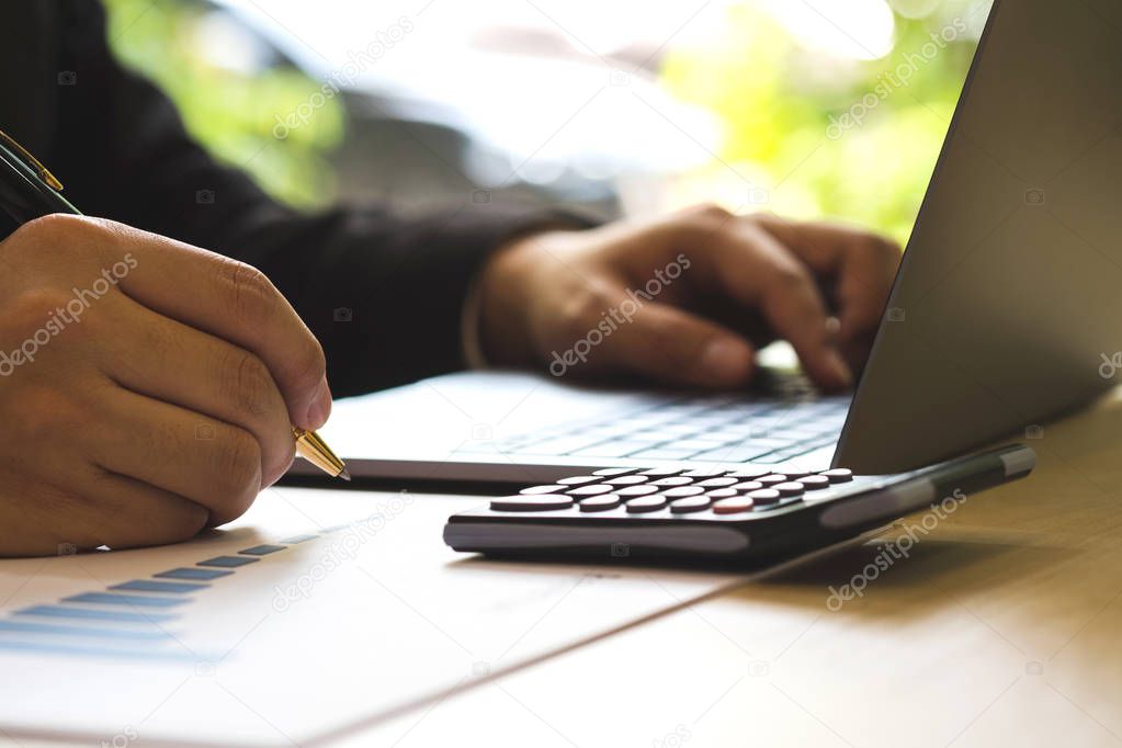 Success business man checking report profit on paper with rising charts, discussing company growth. Close up businessman hand holding pen and taking note in paper and laptop on wood desk in workplace.