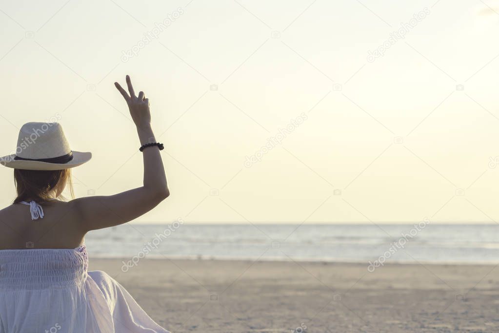 Back side of happy woman sitting on tropical sea and beach showing two fingers or victory sign during sunset. Young woman enjoy life vacation and relaxing in paradise island in vintage tone.