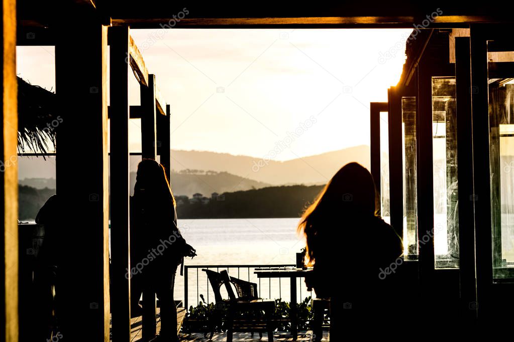 Silhouette of woman walking into cafe side the sea with beautiful blazing sunset landscape at mountain, sea and orange sky background with awesome sun golden reflection. Sunset view at restaurant.