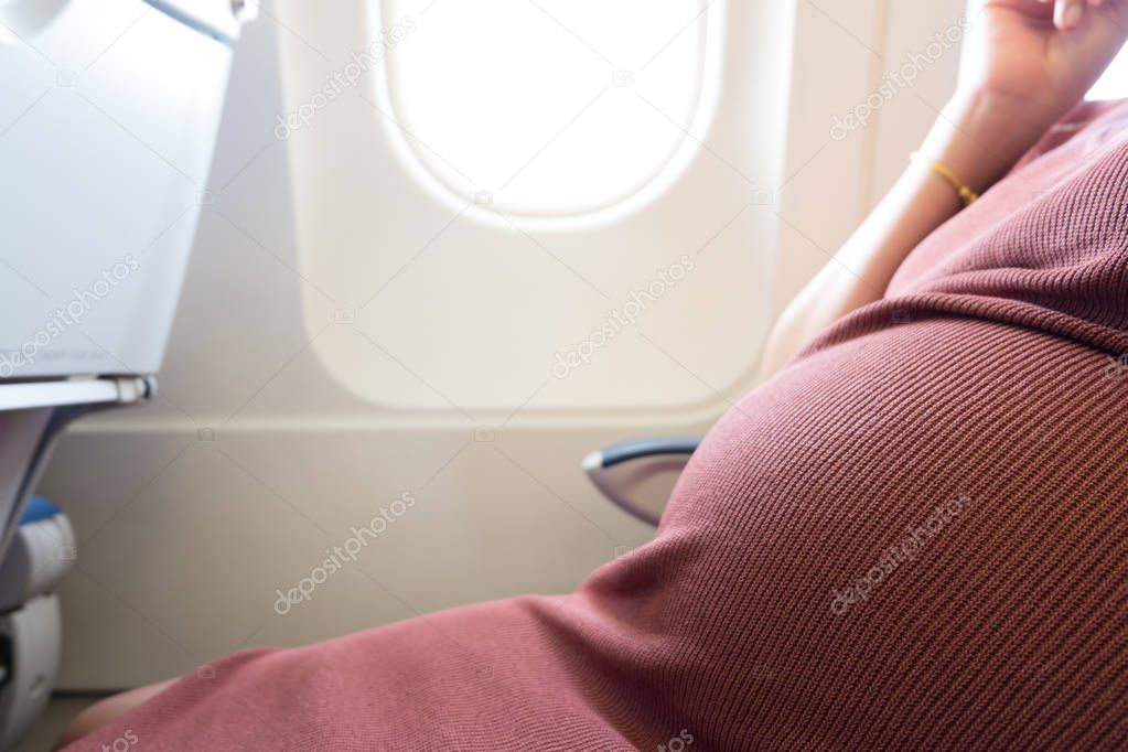 Happy pregnant woman is traveling to destination by plane sitting at window seat light are shining from porthole. Pregnancy healthy and travel concept.