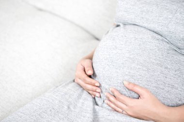 Gri kanepe evde ve karnı üzerinde dokunmadan tutan el üzerinde oturan genç güzel hamile kadın. Mutlu Asyalı hamile kadın aşk ve annelik kavramı kopya alanı ile kanepede rahatlatıcı.