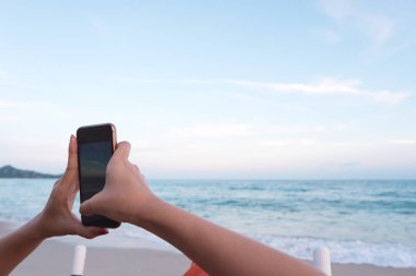 Kadın el deniz görünümü fotoğraf ve video plaj tatil plaj sandalyede otururken alarak smartphone tutarak kapatın. Kadın seyahat keyfini çıkarın tatil tropikal deniz yaz aylarında. Yaşam tarzı, teknoloji.
