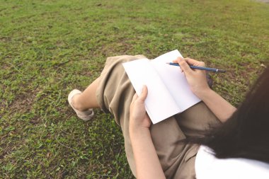 Genç kadın kalem çim açık park üzerinde ile defter üzerinde yazma. Üniversiteli kız çalışmak için bahçede oturan rahat beyaz gömlek, kahverengi pantolon giyer. Yazma fikri, Blogger ve eğitim kavramı.