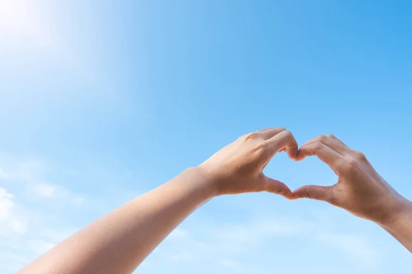 Female hands in heart shape over blue sky background in sunny day. Hands making love sign and symbol. Love, summer travel, nature outdoor holiday and freedom concepts.