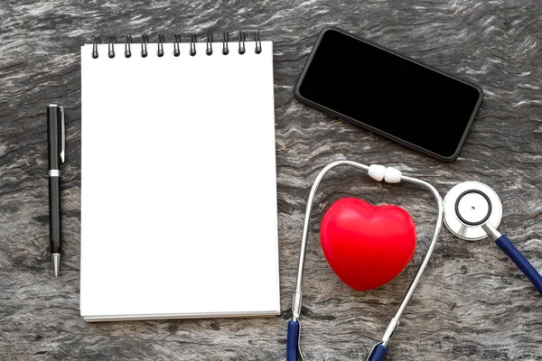 Health red heart with stethoscope and blank notebook for input some text with pen and blank screen of smartphone on wooden background with copy space. World health heart day and daily health concept.