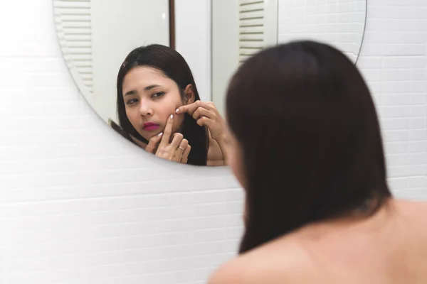 Young asian woman looking at mirror with acne on her face in bathroom. Beautiful girl squeezing her pimple, removing pimple from face. Skin care. Acne spot pressing on skin problem.