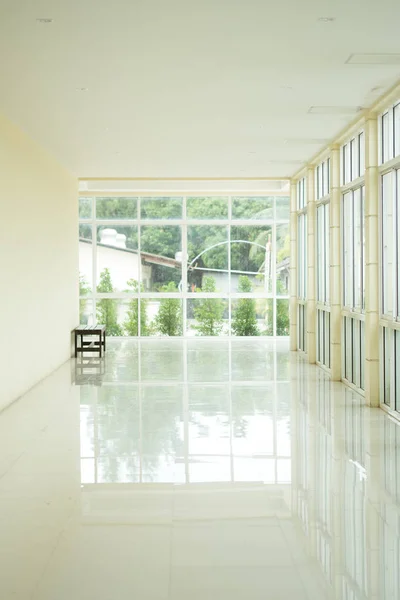 Empty walkway in building hall perspective with long white wall and long windows with light at front.
